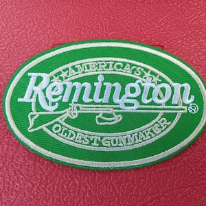 May include: An oval-shaped embroidered patch featuring a green background with a white border. The text reads "AMERICA'S Remington OLDEST GUNMAKER" in white embroidery, accompanied by a white graphic illustration of a long gun centered horizontally.