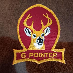 May include: Embroidered patch featuring a deer head with six-point antlers, set against a red oval background with a gold border. The text "6 POINTER" is displayed on a gold banner below the deer. The patch is likely for apparel or accessories.
