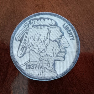 May include: A circular embroidered patch featuring a light gray and white design of a Native American profile, similar to a Buffalo nickel. The patch includes the word LIBERTY printed on the right side and the year 1937 on the lower left side.