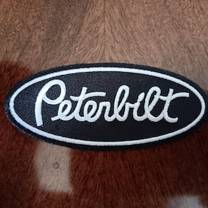 May include: An oval-shaped embroidered patch featuring a black background with a white border and the word Peterbilt stitched in white script lettering across the center. The patch has a textured fabric surface and a clean, defined edge.