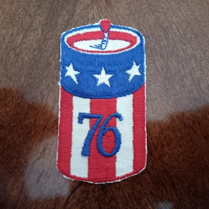 May include: Embroidered patch in the shape of a firecracker, featuring red and white stripes, a blue top with white stars, and the number "76" in blue. The patch has a red fuse and is likely a patriotic item.