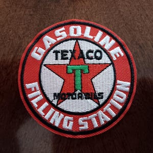 May include: A circular embroidered patch featuring a red border with the white text GASOLINE FILLING STATION. The center displays a white circle with a red star, a green letter T, and the black text TEXACO MOTOR OILS.