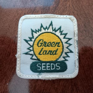 May include: A square patch with a light blue background and a white border. The patch features a yellow sunburst design with the words "Green Land" in green script. Below the sunburst, the word "SEEDS" is displayed in green.