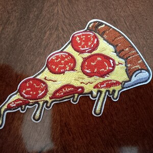 May include: Embroidered pizza slice patch with a white border. The pizza has a yellow cheese base, red pepperoni, and brown crust. Melted cheese drips down the side, adding a realistic touch to the design.