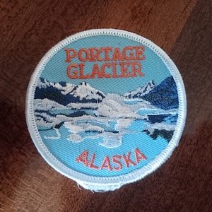 May include: A blue and white round embroidered patch with the text "Portage Glacier Alaska" in orange thread. The patch features a scenic image of a glacier with snow-capped mountains in the background.