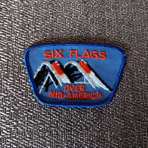 May include: A blue patch with the words "SIX FLAGS OVER MID-AMERICA" in orange and white. The patch features a mountain design with red and white accents. The patch is likely a souvenir or collectible item.
