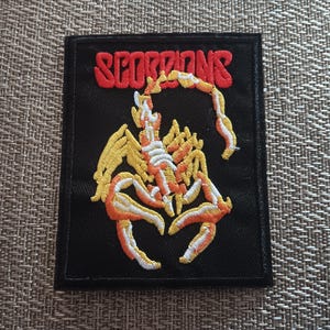 May include: A rectangular black embroidered patch featuring the word SCORPIONS in red capital letters at the top. Below the text is a detailed yellow and orange scorpion graphic with white highlights, all stitched onto a black fabric background.