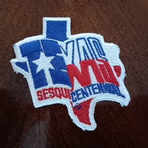 May include: A white, blue, and red embroidered patch shaped like the state of Texas. The patch features the word "TEXAS" in red and blue lettering and the words "SESQUICENTENNIAL" in blue lettering.