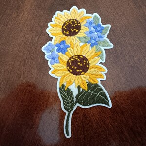 May include: Embroidered patch featuring two sunflowers with yellow petals and brown centers, accompanied by blue hydrangea flowers and green leaves. The patch has a white border and is likely for clothing or accessories.