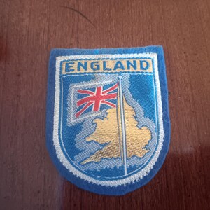 May include: A blue felt patch with white stitching and the word "ENGLAND" in white letters. The patch features a gold outline of the United Kingdom with a red, white, and blue flag.
