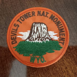 May include: Orange embroidered patch with the text "Devils Tower Nat Monument WYO". The patch features a brown and white embroidered image of Devils Tower National Monument.