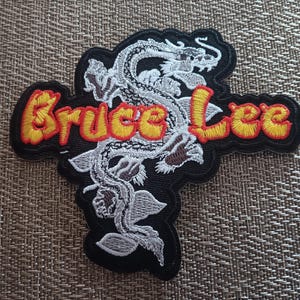May include: A black embroidered patch with a white dragon and the text "Bruce Lee" in yellow with red outlines.