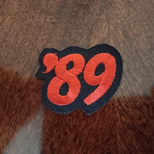 May include: A black embroidered patch with the number "89" in red thread. The patch has a heart-like shape and is set against a brown and gray patterned background.
