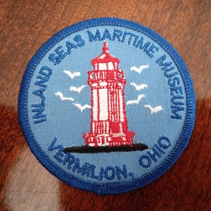 May include: A circular embroidered patch with a light blue background featuring a red and white lighthouse in the center, surrounded by white birds. The text reads Inland Seas Maritime Museum and Vermilion, Ohio in dark blue embroidery.