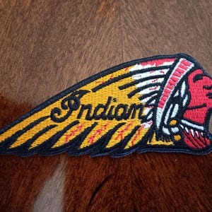 May include: An embroidered patch featuring the Indian Motorcycle logo. The design depicts a profile of a person wearing a red headdress with white and yellow feathers. The word Indian is stitched in black script across the yellow feathers.