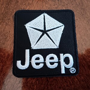 May include: Black square patch with a white pentagon star design and the word "Jeep" in white lettering. The patch has a stitched border and a registered trademark symbol.