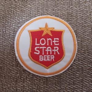 May include: A circular patch with a white background and an orange border. The center features a red shield with the words "LONE STAR BEER" in white lettering, topped with a gold star.