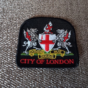 May include: Black patch featuring the City of London coat of arms. The design includes a shield with a red cross, supported by two silver dragons with red accents. The text "CITY OF LONDON" is in red.