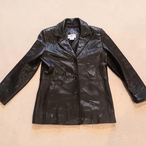 May include: A black leather blazer with a notched collar and three-button closure. The jacket has long sleeves and a tailored fit. A small label is visible near the collar. This classic jacket is suitable for various occasions.