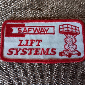 May include: Rectangular white patch with red embroidered text and design. The text reads "SAFWAY LIFT SYSTEMS". A red border frames the patch. The design includes a lift platform illustration. Ideal for workwear or vintage collection.