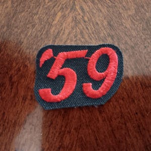 May include: A rectangular fabric patch with rounded corners featuring the embroidered number 59 in bright red thread. The background of the patch is a dark navy blue fabric with a visible woven texture, and the red numbers are stitched in a bold font.