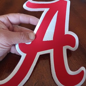 May include: A large, red, fabric letter A patch with a thick white border. The letter features a classic serif typeface design with a textured surface, suitable for application onto various textile items as a decorative or team-themed emblem.