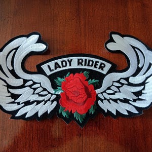 May include: Embroidered patch featuring a red rose and green leaves at the center, flanked by white wings with black outlines. The patch has the text "LADY RIDER" on a black banner. The patch is on a dark wood surface.