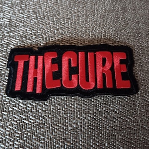 May include: A black patch with the red text "THE CURE". The text is in a bold, sans-serif font. The patch has a black border and is rectangular in shape. The background is a textured, gray fabric.