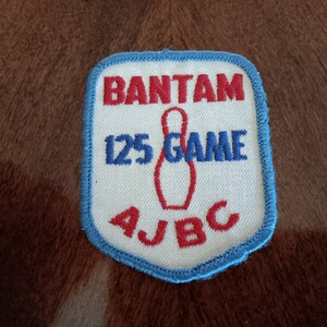 May include: A white patch with blue edging, featuring red embroidered text that reads "BANTAM 125 GAME AJBC". A red bowling pin is embroidered in the center.