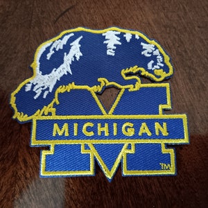 May include: A blue and yellow embroidered patch featuring the word "MICHIGAN" in yellow block letters. Above the word is a blue bear with white detailing. The patch has a yellow border.