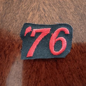 May include: A black fabric patch with the number "76" embroidered in red thread. The patch has a slightly irregular shape and is set against a wooden surface.