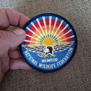 May include: Embroidered patch with a blue border featuring a stylized eagle and sunburst design. The text "NATIONAL WILDLIFE FEDERATION" and "MEMBER" are displayed on the patch. The patch is round and has a detailed design.