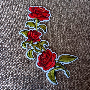 May include: Embroidered patch featuring a floral design of three red roses with green leaves and stems. The patch has a white border and is set against a textured brown background. Ideal for adding to clothing or accessories.