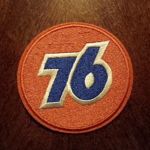 May include: Embroidered patch featuring the number "76" in blue with a white outline, set against an orange background. The patch is circular and has a textured appearance, likely for application on clothing or accessories.