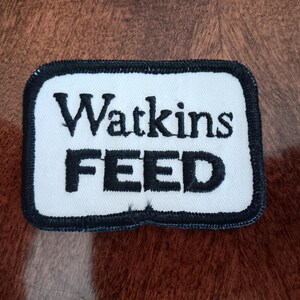 May include: A rectangular patch with the words "Watkins FEED" in black lettering on a white background. The patch has a black border and is set against a brown background.