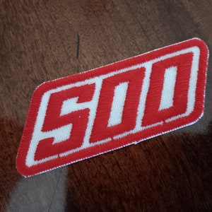 May include: Embroidered patch with the number "500" in red, outlined in white, on a white background. The patch is rectangular with rounded corners. The patch is likely for a jacket or bag.