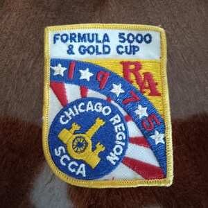May include: A rectangular embroidered patch featuring the text FORMULA 5000 & GOLD CUP at the top. The design includes a blue arc with white stars, the year 1975, and a circular emblem with the text CHICAGO REGION SCCA and a yellow cannon icon.