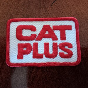 May include: Embroidered patch with the words "CAT PLUS" in red, stitched onto a white background. The patch has a red border and a rectangular shape. The patch is placed on a brown surface.