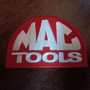 May include: A red, semi-circular patch with the embroidered text "MAC TOOLS" in white. The patch has a white border and is likely for application on clothing or equipment.