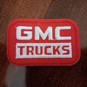 May include: A rectangular red patch with white embroidered text. The top line reads "GMC" and the bottom line reads "TRUCKS". The patch has a white border and rounded corners. The background is a textured brown surface.