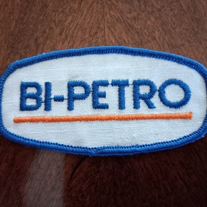 May include: A white oval patch with blue edging and the text "BI-PETRO" in blue embroidery. A thin orange line is embroidered below the text.