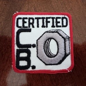May include: Embroidered patch with a white background, red border, and the words "CERTIFIED C.B." in black. A gray, stylized nut graphic is on the right side of the patch. The patch is square-shaped.
