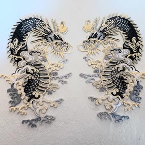 May include: Two symmetrical embroidered patches featuring mirror-image dragons in black, white, and cream thread. The dragons are depicted with long, winding bodies, detailed scales, and flowing cloud or water motifs surrounding their lower sections.