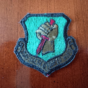 May include: A green and gold embroidered patch with a fist holding a knife. The patch has the text "Special Forces" around the edge.