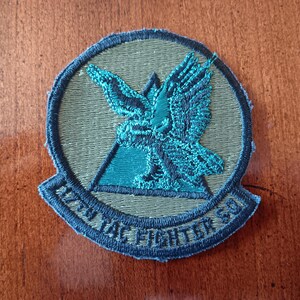 May include: A green and blue embroidered patch with a stylized eagle and a triangle. The text "27th TAC Fighter Sq" is embroidered around the edge.