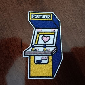 May include: A white, yellow, and blue embroidered patch of an arcade game with the text "GAME ON" on the top. The game has a pink heart in the screen.