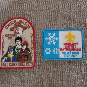 May include: Two vintage Boy Scout patches. One is a red, white, and tan patch with a portrait of a man in a hat and the text "Chuck Mason District Pioneer Fall Camporee 1979." The other is a blue and white patch with snowflakes and the text "Sinnissippi District Winter Camporee Valley Forge 1777-1977."