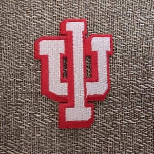 May include: Embroidered patch featuring the Indiana University logo, a stylized "IU" in white with a red border. The patch is rectangular and likely designed to be sewn or ironed onto fabric. The background is a textured, neutral-toned surface.