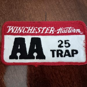 May include: Rectangular patch with a red border and the text "WINCHESTER-Western" in white. The patch has a white center with the black letters "AA" and the numbers "25 TRAP".