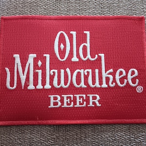 May include: Red fabric patch with white embroidered text that reads "Old Milwaukee Beer" with a registered trademark symbol.